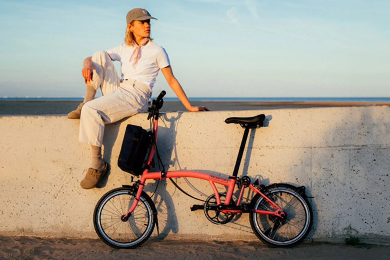 A person sitting on a wall with a flaming pink Bromtpon bicycle by their side, against a scenic background. 