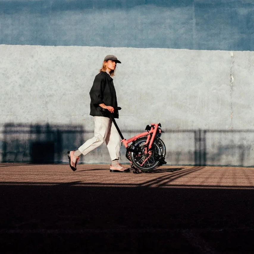 A person walking with a flaming pink Brompton folded bike on a dock.