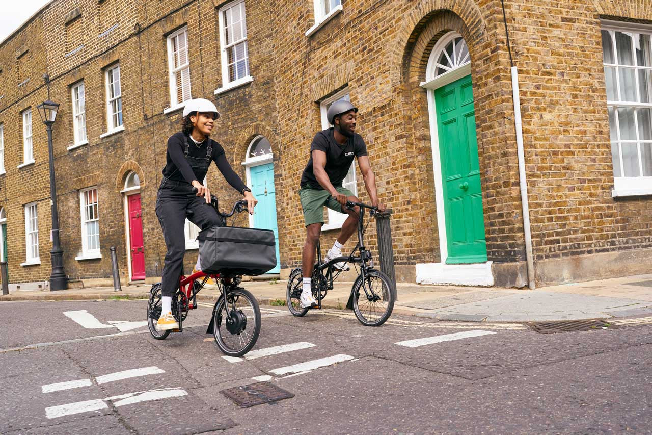 Two people riding Brompton folding bikes on a city street with brick buildings in the background.
