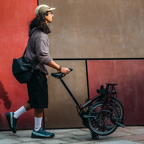 A person with a backpack pushing a Brompton folded bike against a red wall.