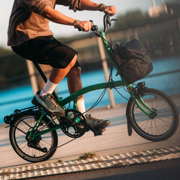 A person riding a green Brompton folding bicycle by a water.