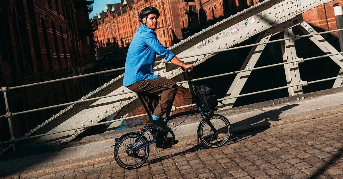 A person riding Brompton Electric folding bike on the bridge in a city setting.