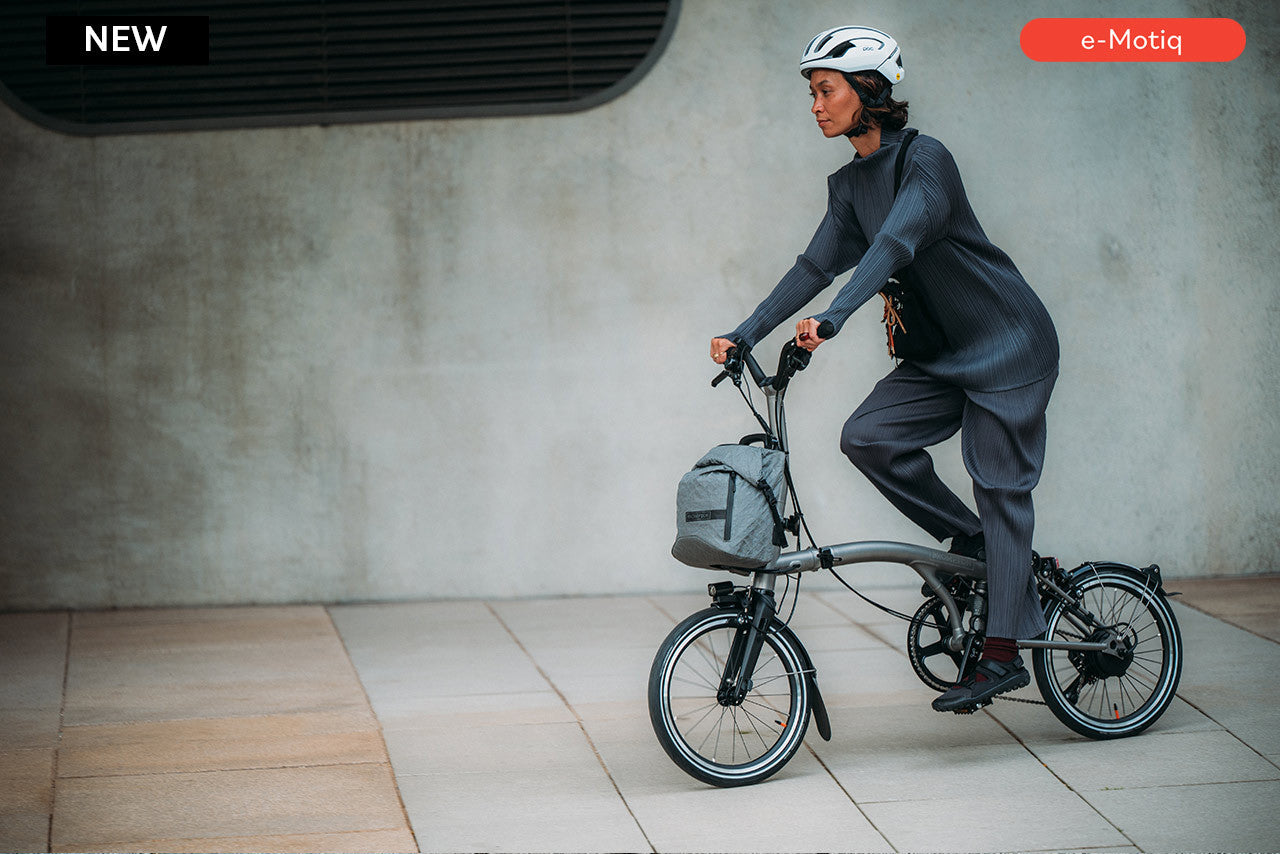 Person riding a Brompton Electric T Line bicycle with a backpack in an urban setting, featuring e-Motiq branding.