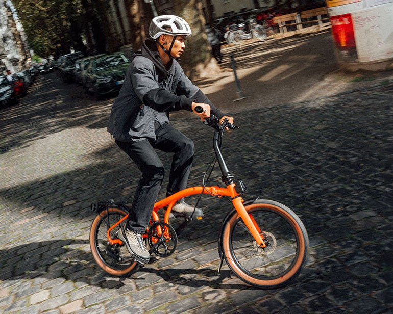 A person riding an orange Brompton G Line bicycle on a city street