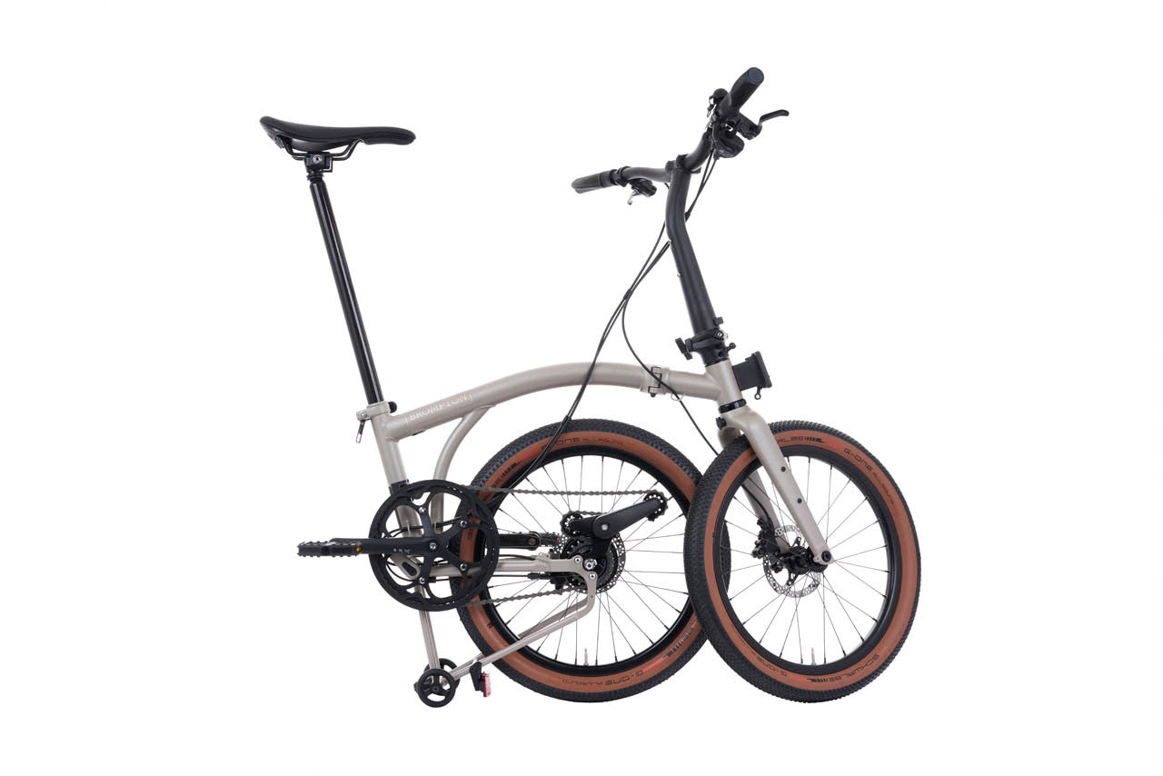 A compact folding bicycle with a light gray frame, black components, and brown sidewall tires, shown on a plain white background.