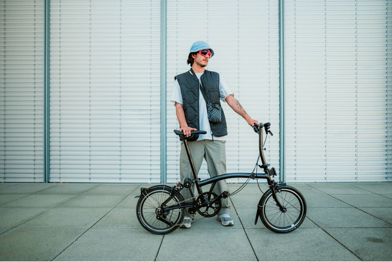 Person holding a Brompton folding bicycle against a light-colored wall.