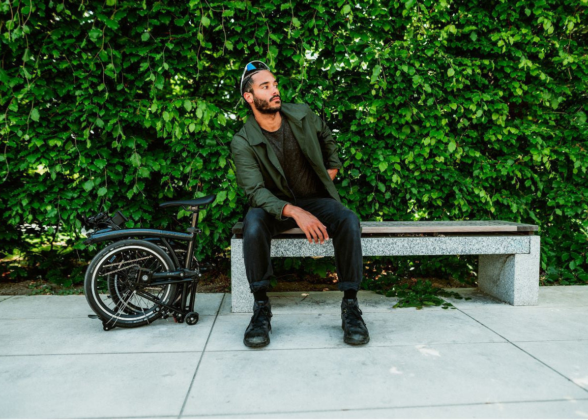 Man sitting on a bench with a folded bike next to him against a green hedge background