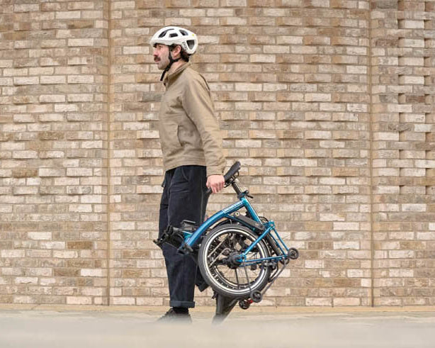 Man carrying a folded Brompton bicycle against a brick wall
