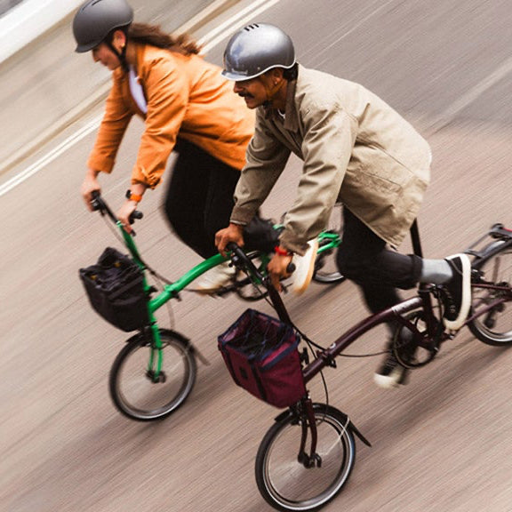 Two people riding Brompton folding bicycles on a road, one with a green bike and the other with a dark purple.