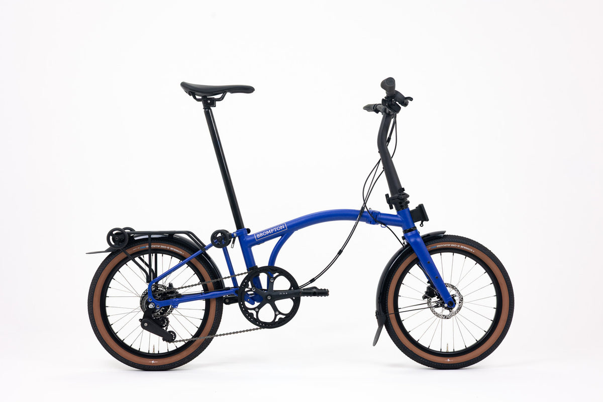 A blue folding Brompton bicycle with black components and brown sidewall tires, shown from a side profile on a clean white background.