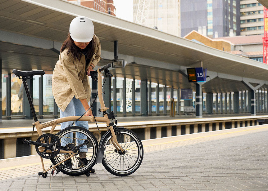 Person unfolding a Brompton folding bike on a platform with a cityscape background.