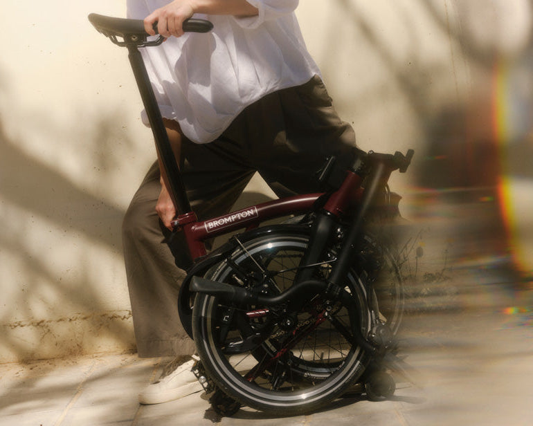Person holding a folded bicycle against a light-colored wall.