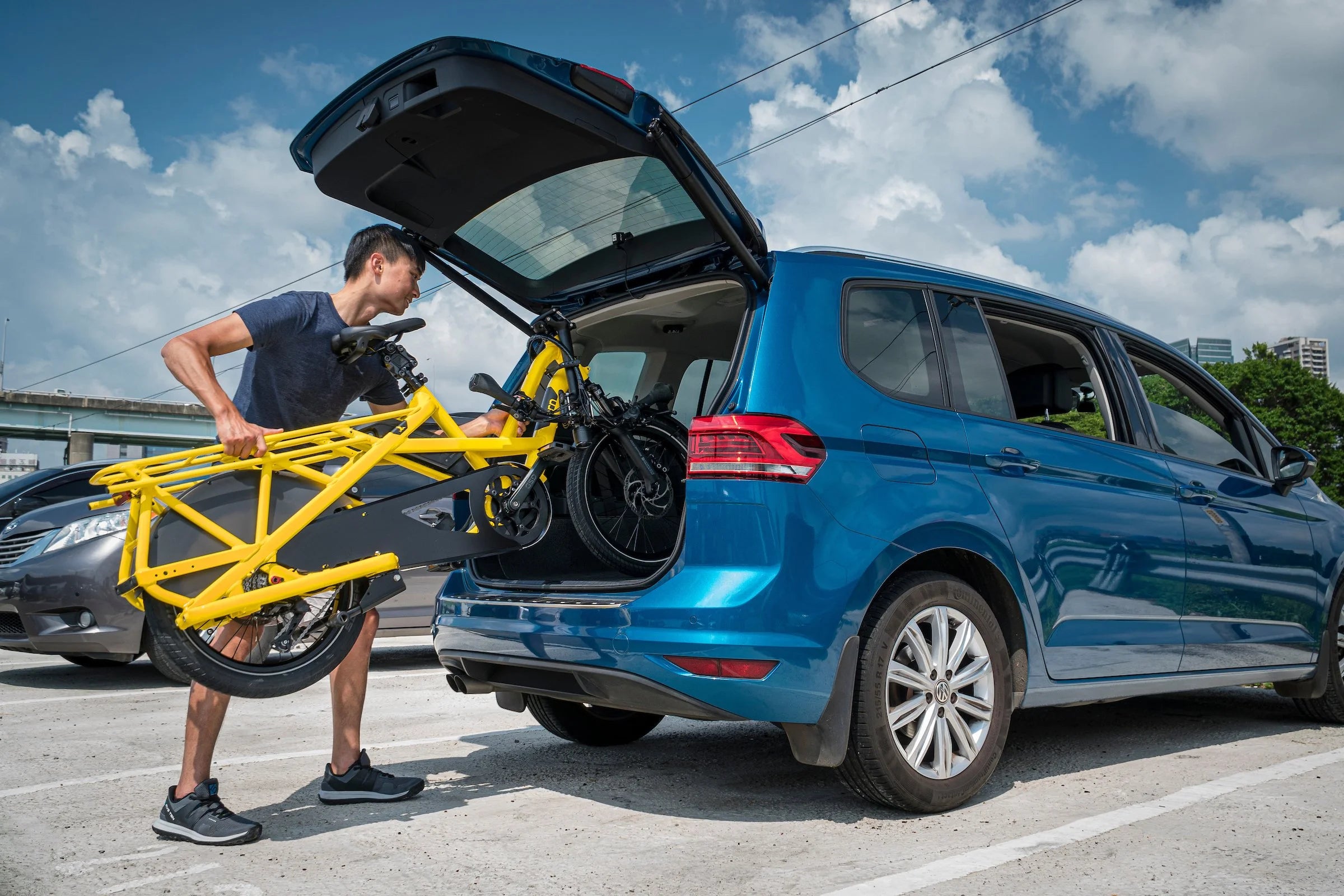 Person loading a yellow bike into the trunk of a blue car on a sunny day.