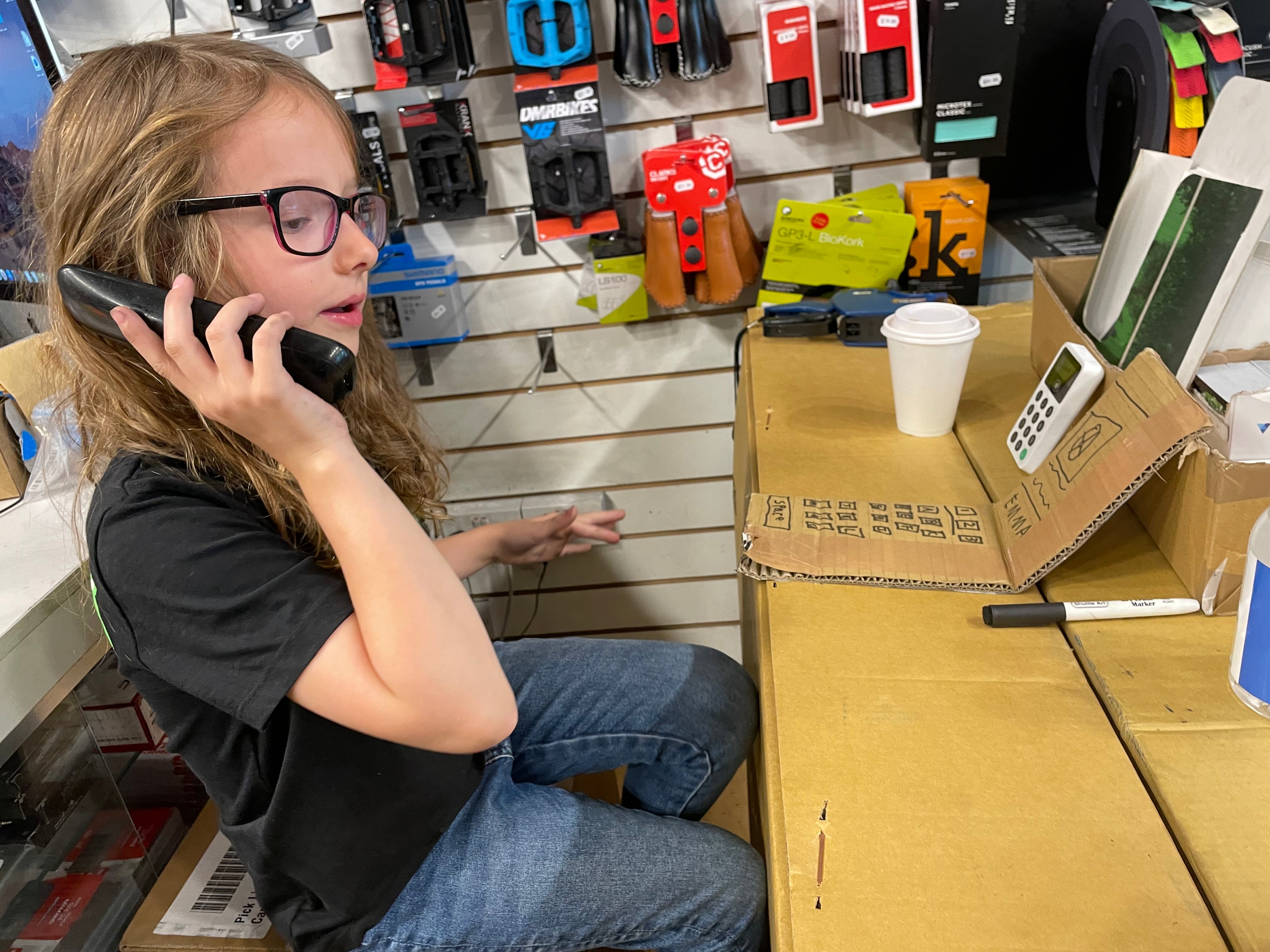 Person sitting at a counter in a store, talking on a phone with various items and products in the background.