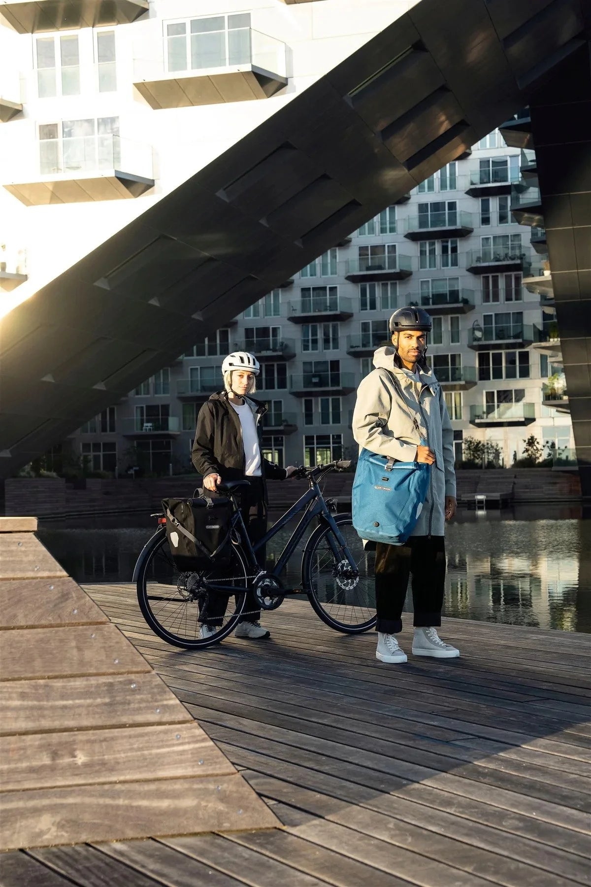 Two people with bicycles on a wooden platform with modern buildings in the background