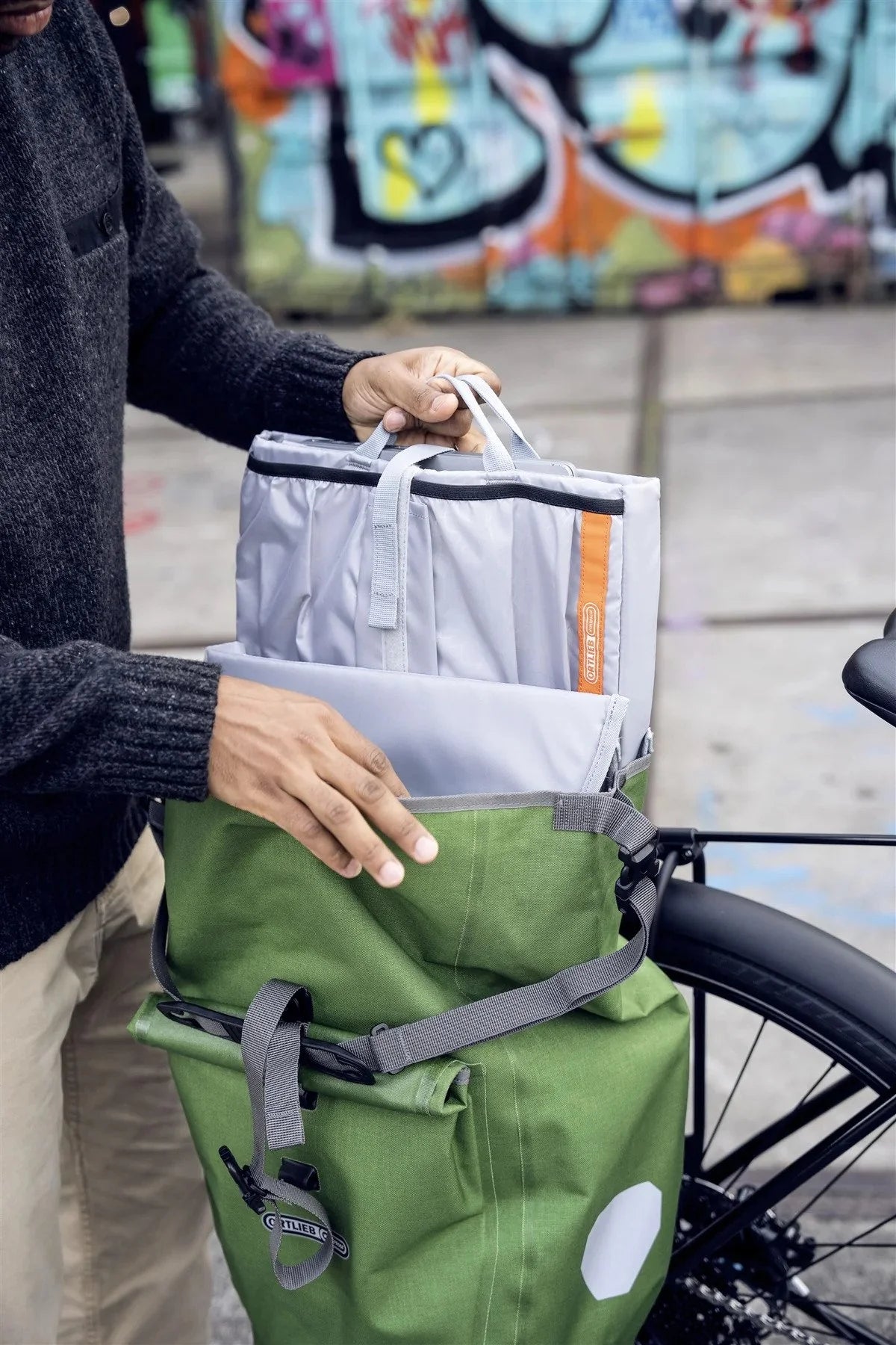 Person holding a green and gray pannier bag with a bicycle in the background and graffiti on the wall.