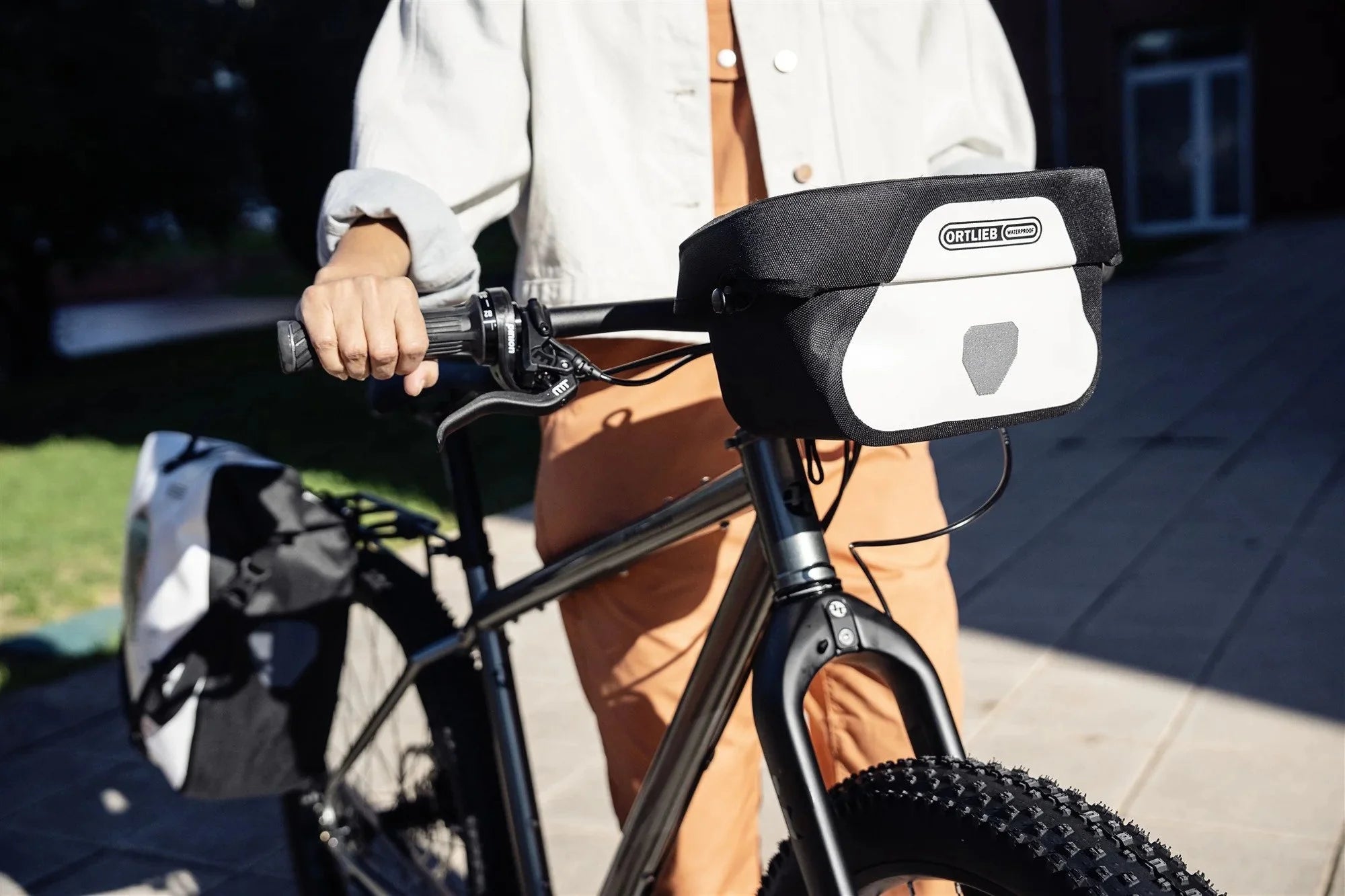 Person holding a bicycle with a white and black Ortlieb handlebar bag attached to the handlebar.