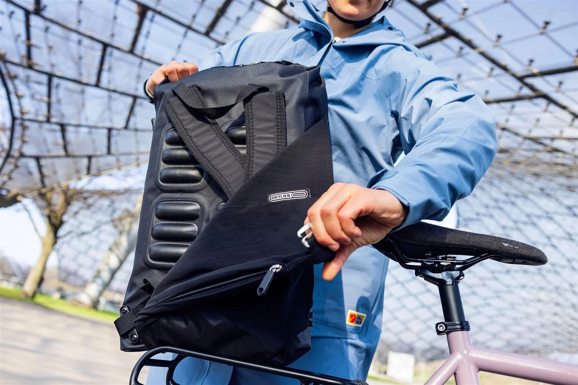 Person holding a black backpack with a brand logo, standing next to a bicycle outdoors.