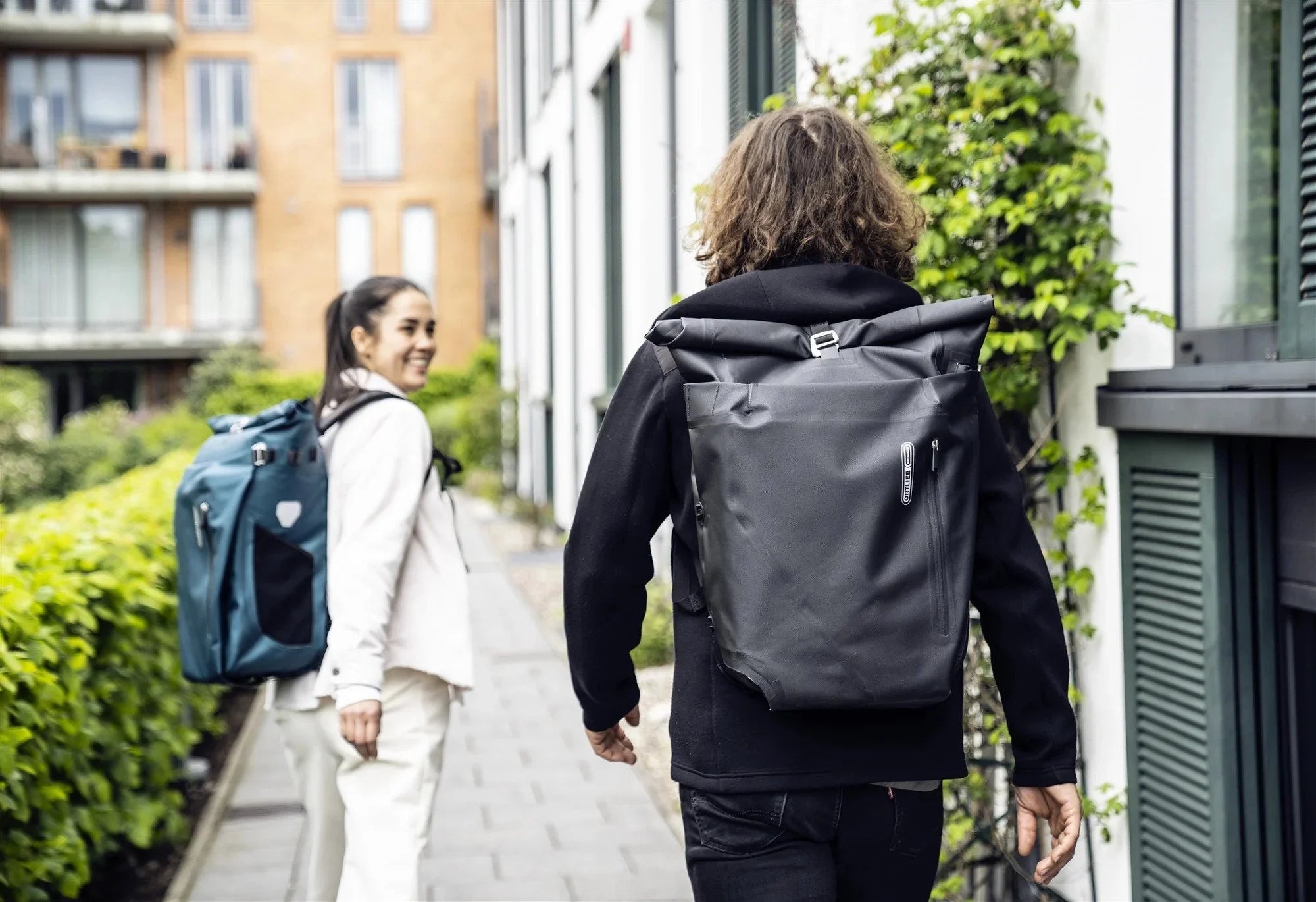 Two people walking outdoors with backpacks, one in a city setting.