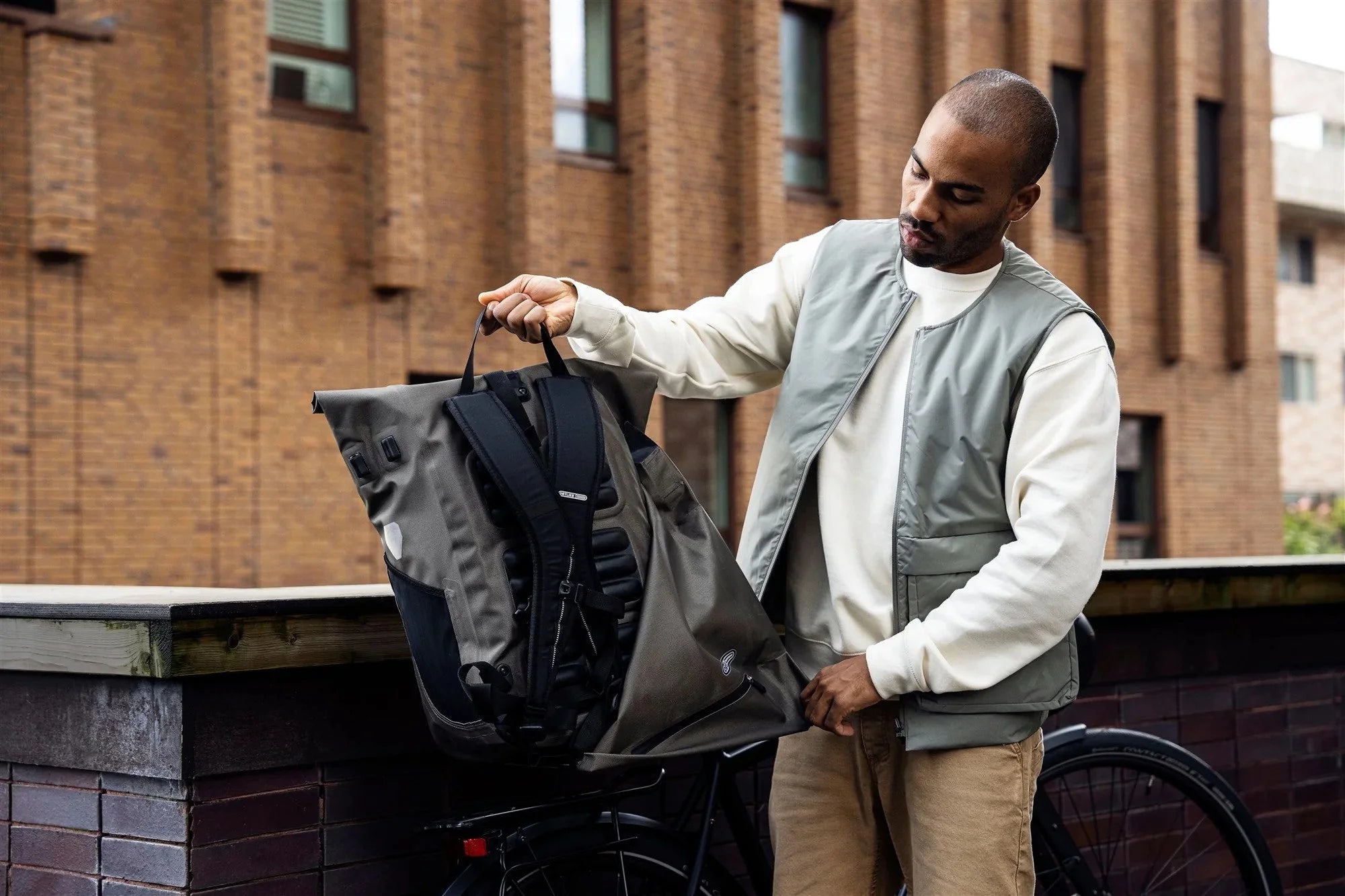 Man in a vest and white shirt looking at a backpack on a bicycle in an urban setting.