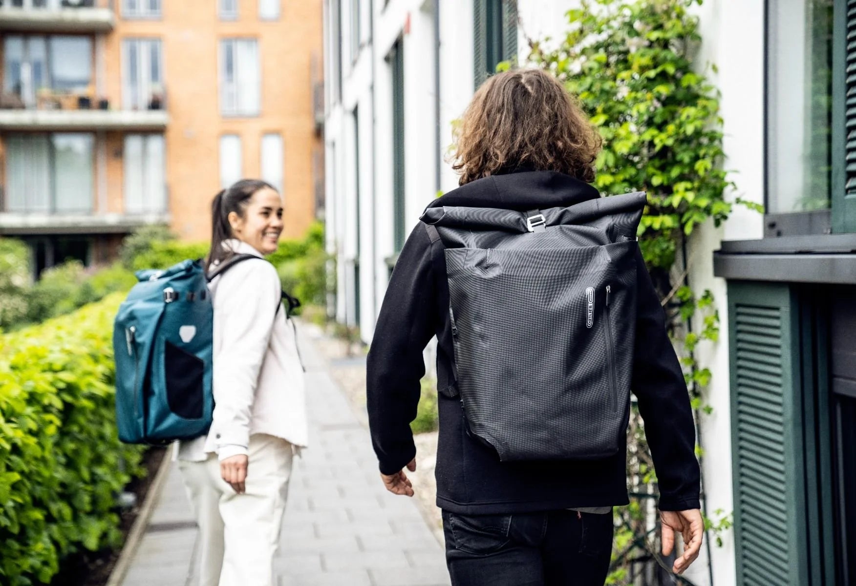 Two people walking outdoors, one with a black backpack and the other with a blue backpack.