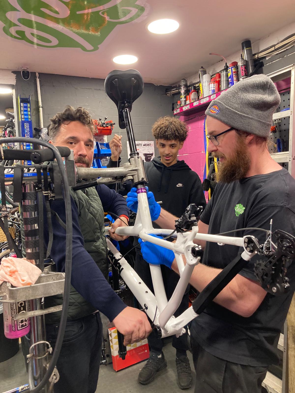 Three men working on a bicycle frame inside a bike shop.