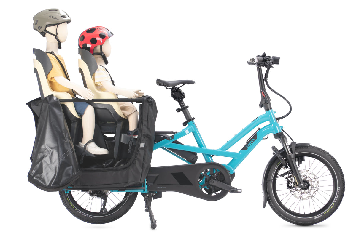 A turquoise and black Tern cargo bike with a front cargo box carrying two mannequin figures wearing helmets, photographed on a clean white background.