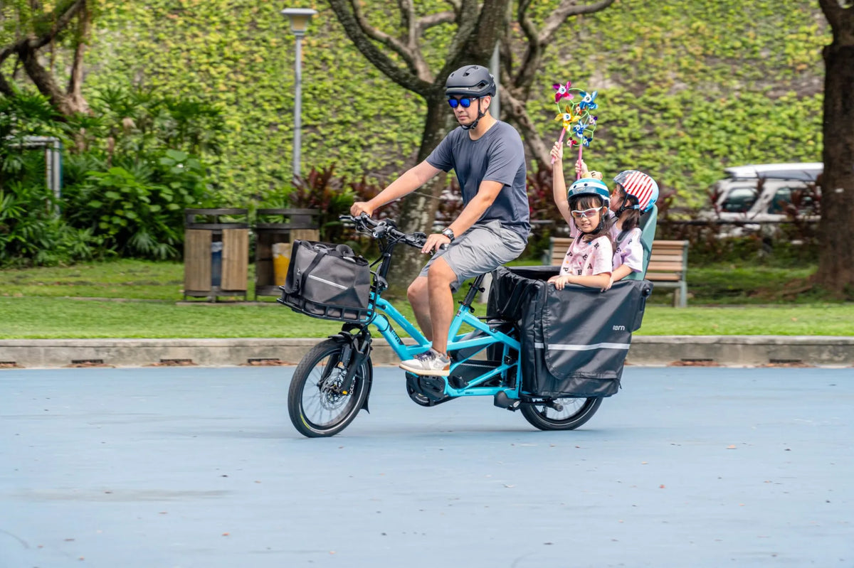 A person riding on the blue and black Tern cargo electric bike with two children wearing helmets in the park setting