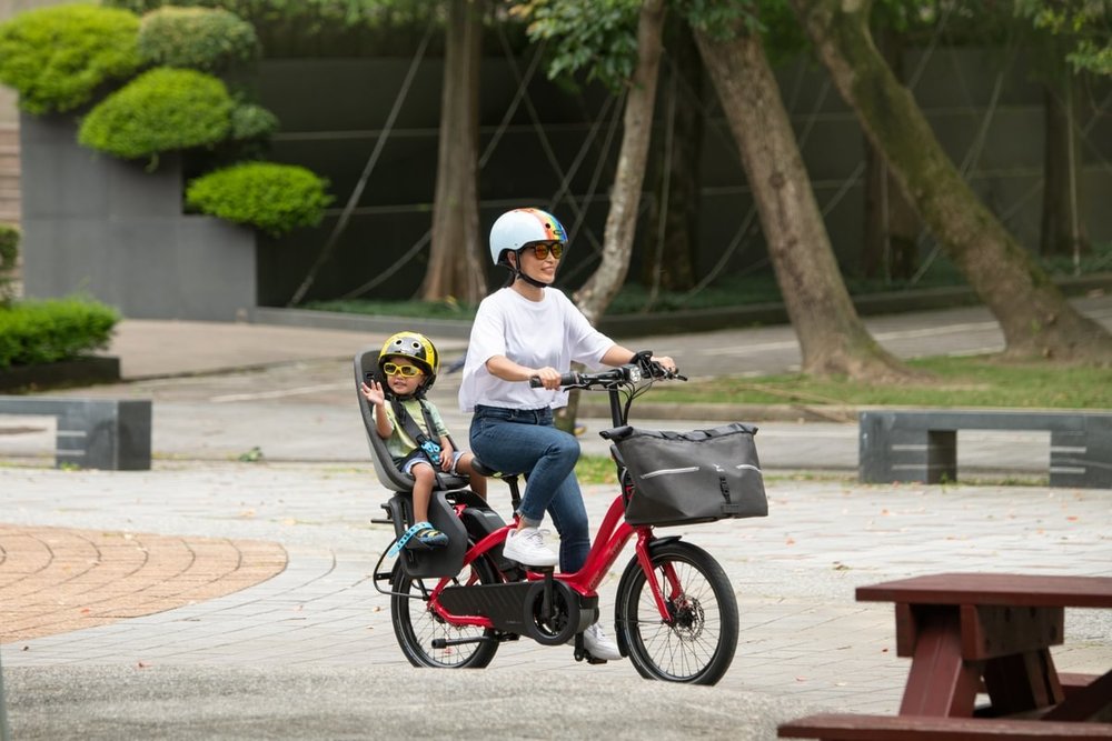 Woman riding a red electric bike with a child in a seat, surrounded by trees and a bench.