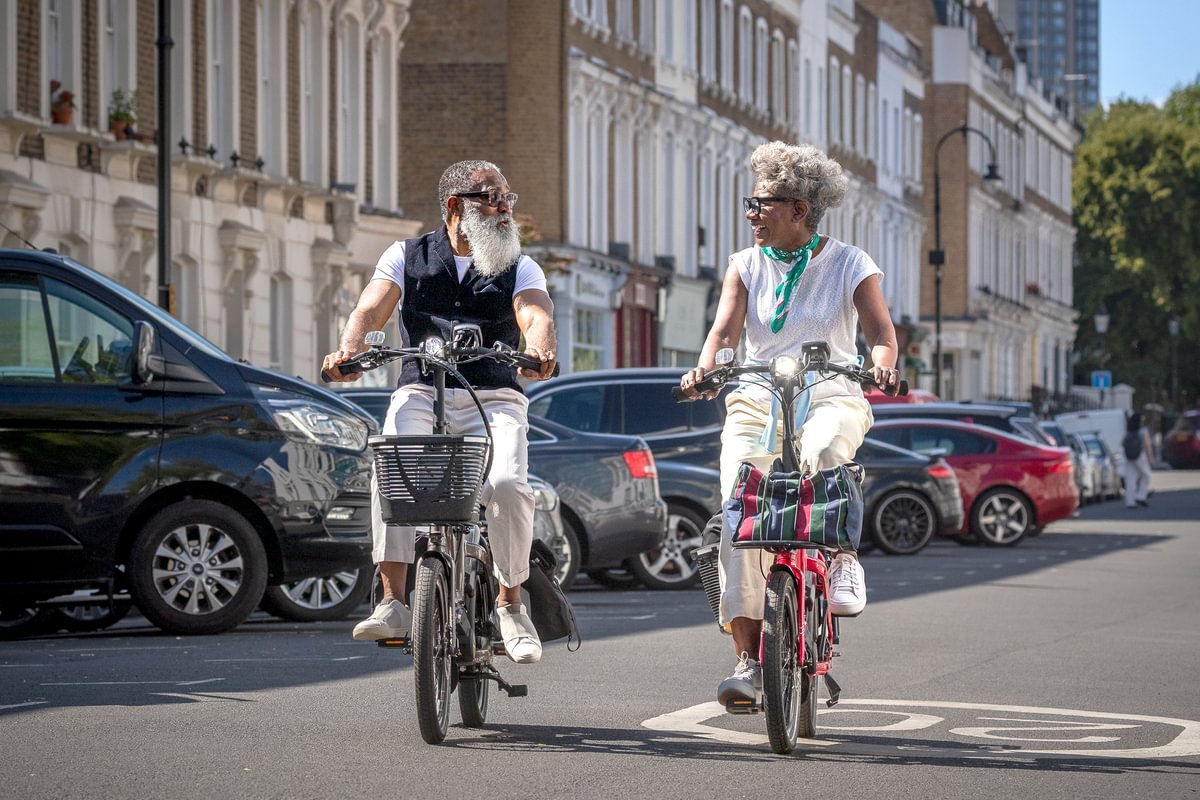 Two people riding bicycles on a city street with cars and buildings in the background.