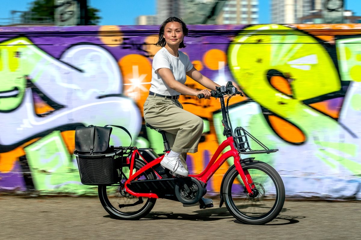 Person riding a red electric bike with a basket in front of a colorful graffiti wall.