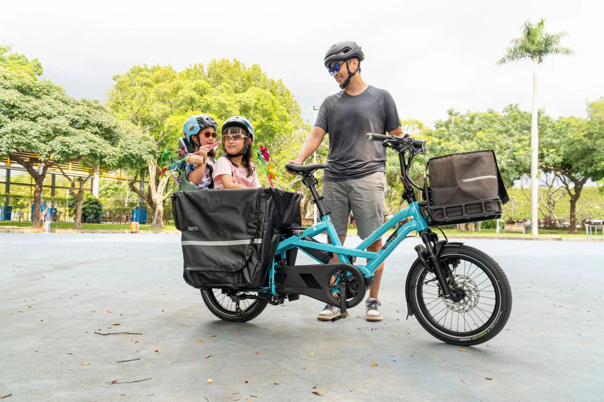 A person standing next to a blue and black Tern cargo bicycle with two kids setting at the back in the park setting.