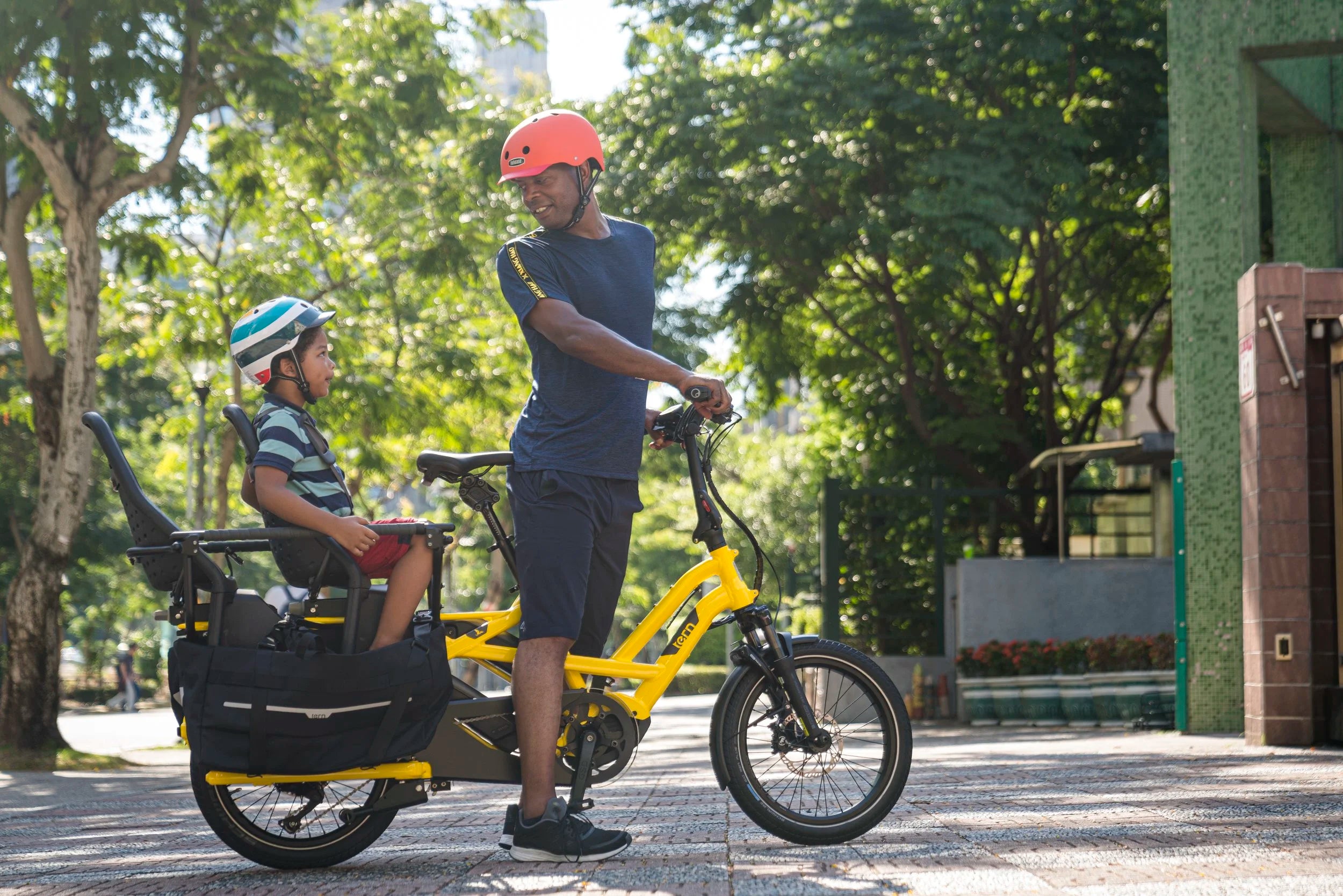 Two people on a yellow cargo bike in an urban setting with trees and buildings in the background.