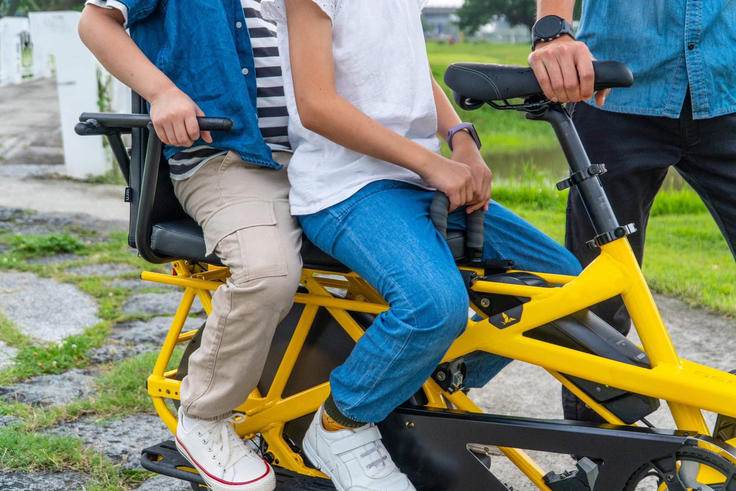 Two people riding a yellow cargo bicycle on a path with grass and a white wall in the background.