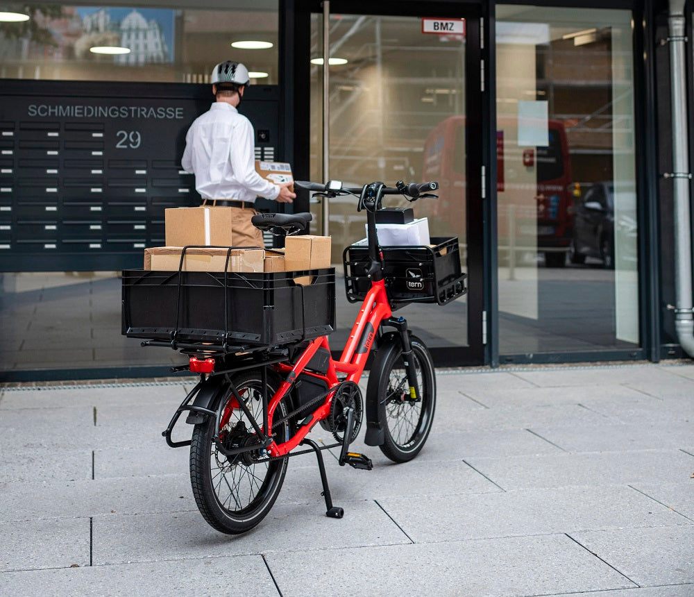 Red Tern cargo bike with a black basket carrying packages, parked outside a building.