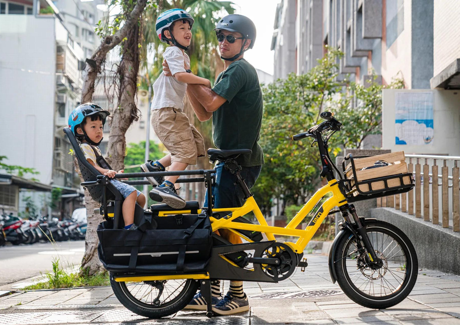 Person riding a yellow cargo bike with two children in front, on a city street.
