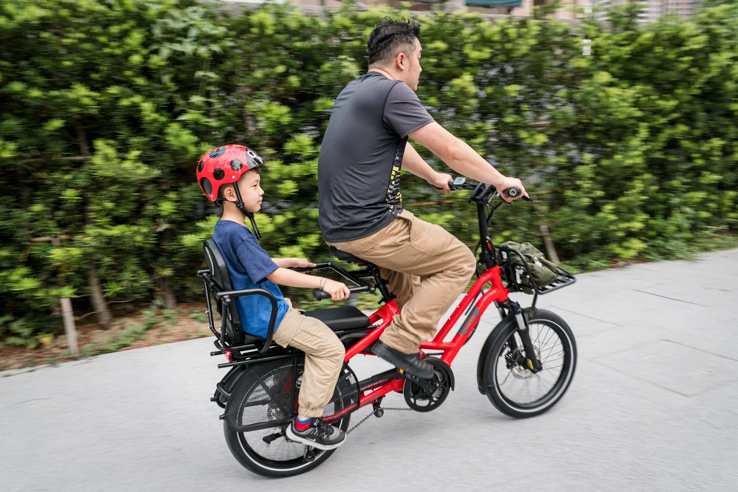 Man and child riding a red electric bike on a path with greenery in the background