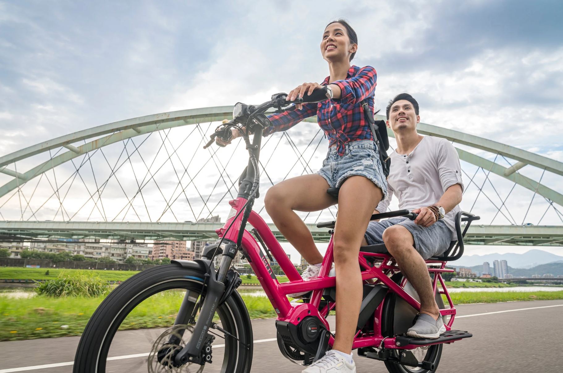 Two people riding a pink electric bike with a bridge in the background