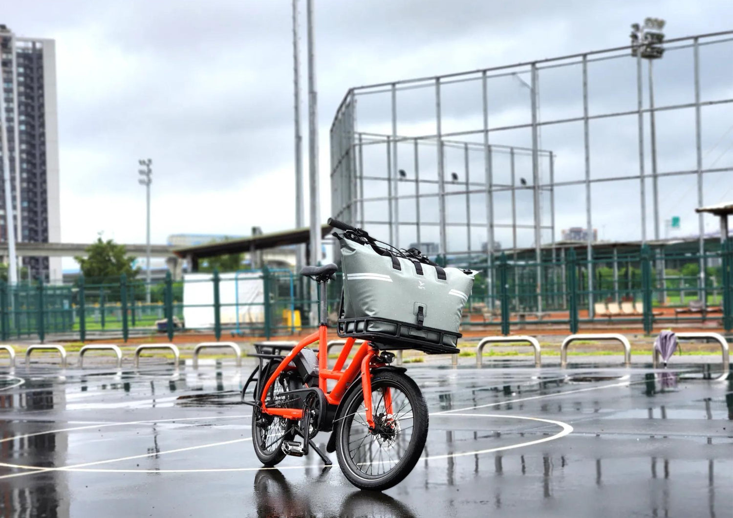 Orange electric bike with a basket on a wet pavement, with a modern building in the background.