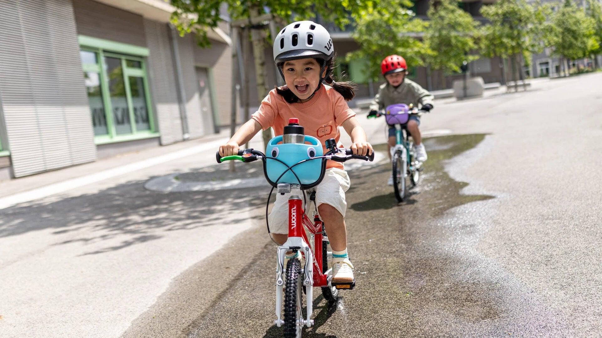 Two children riding bicycles on a city street with trees and buildings in the background.