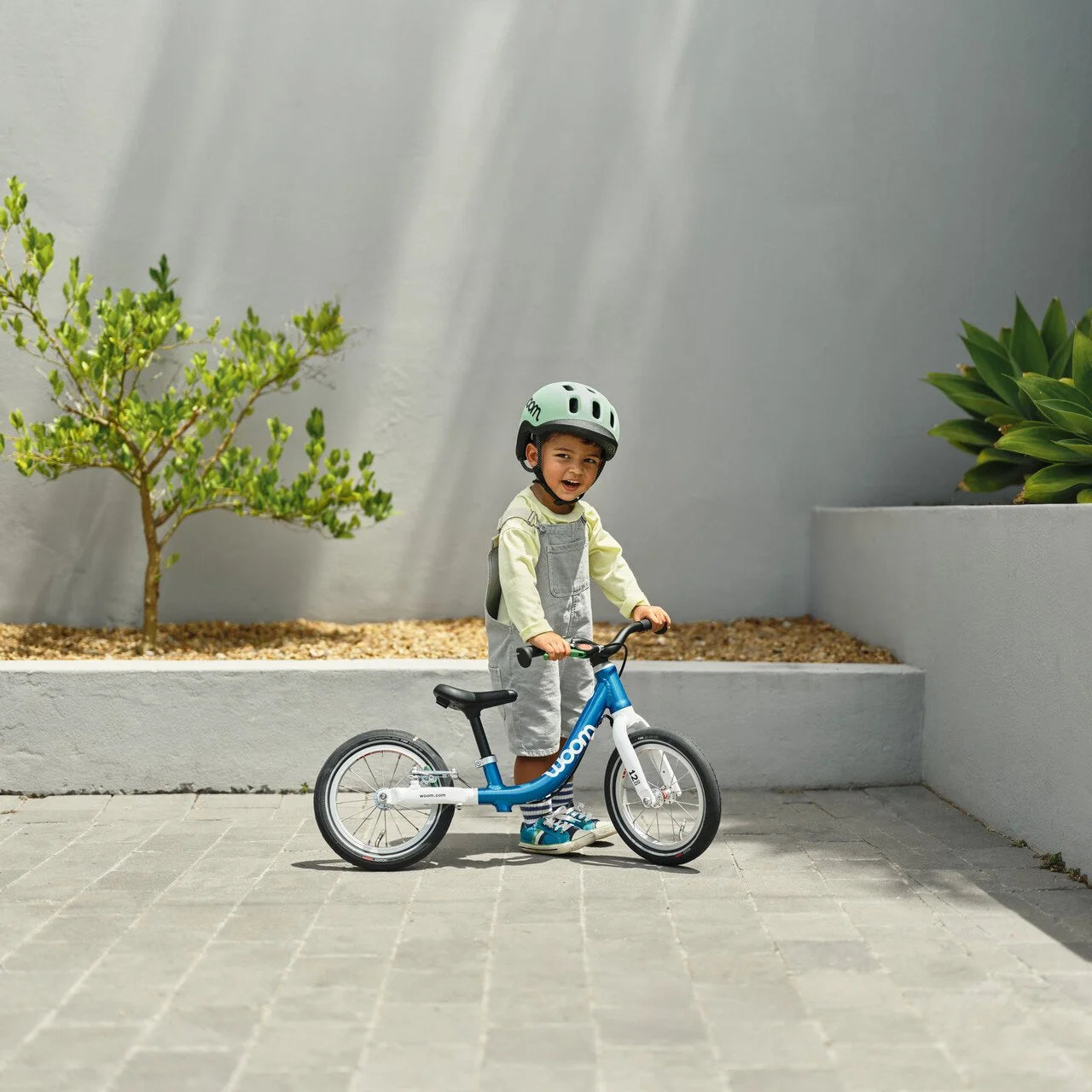 Child standing with a blue Woom balance bike outdoors with a grey wall and plants in the background.