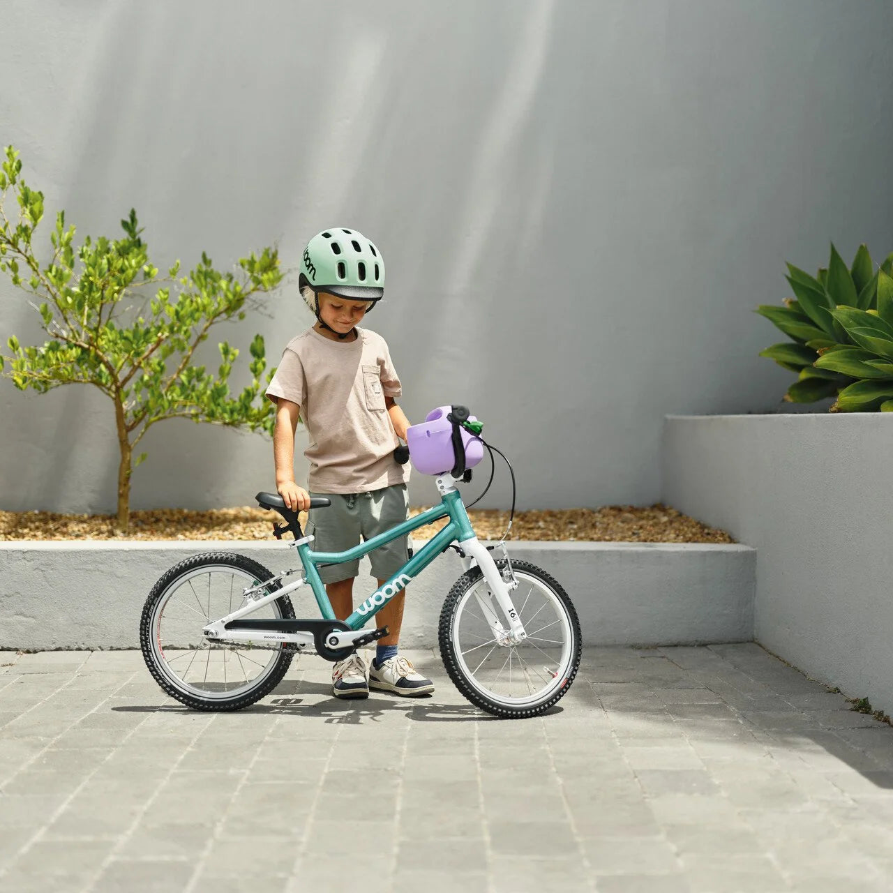 Child with a teal bicycle and helmet standing on a paved area with plants in the background
