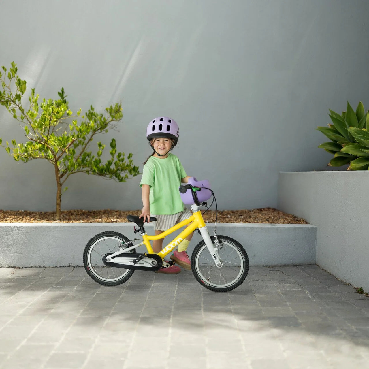 Child with a yellow Woom balance bike and helmet standing on a tiled floor with plants in the background.