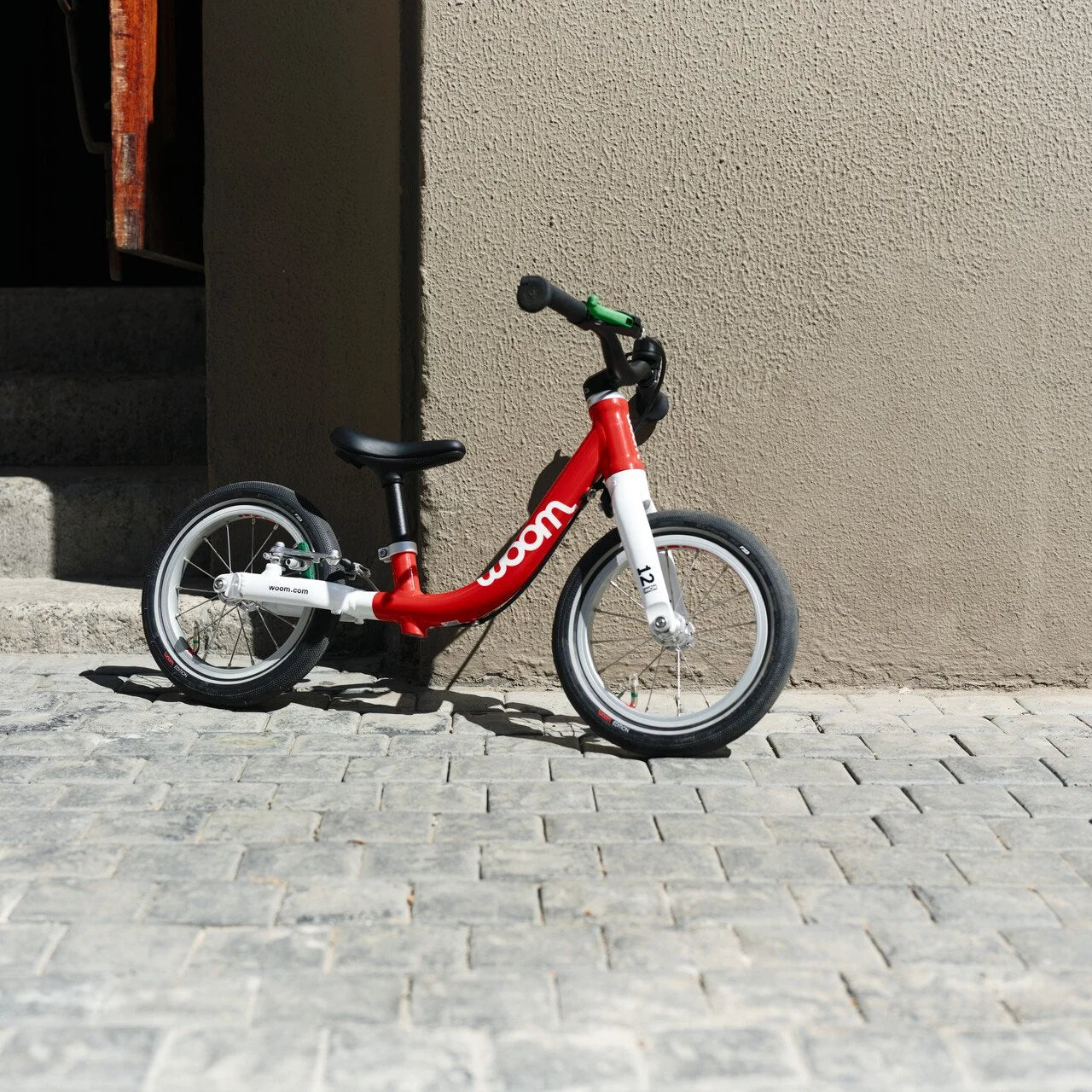 Red and white Woom balance bike on a paved surface with a beige wall in the background.