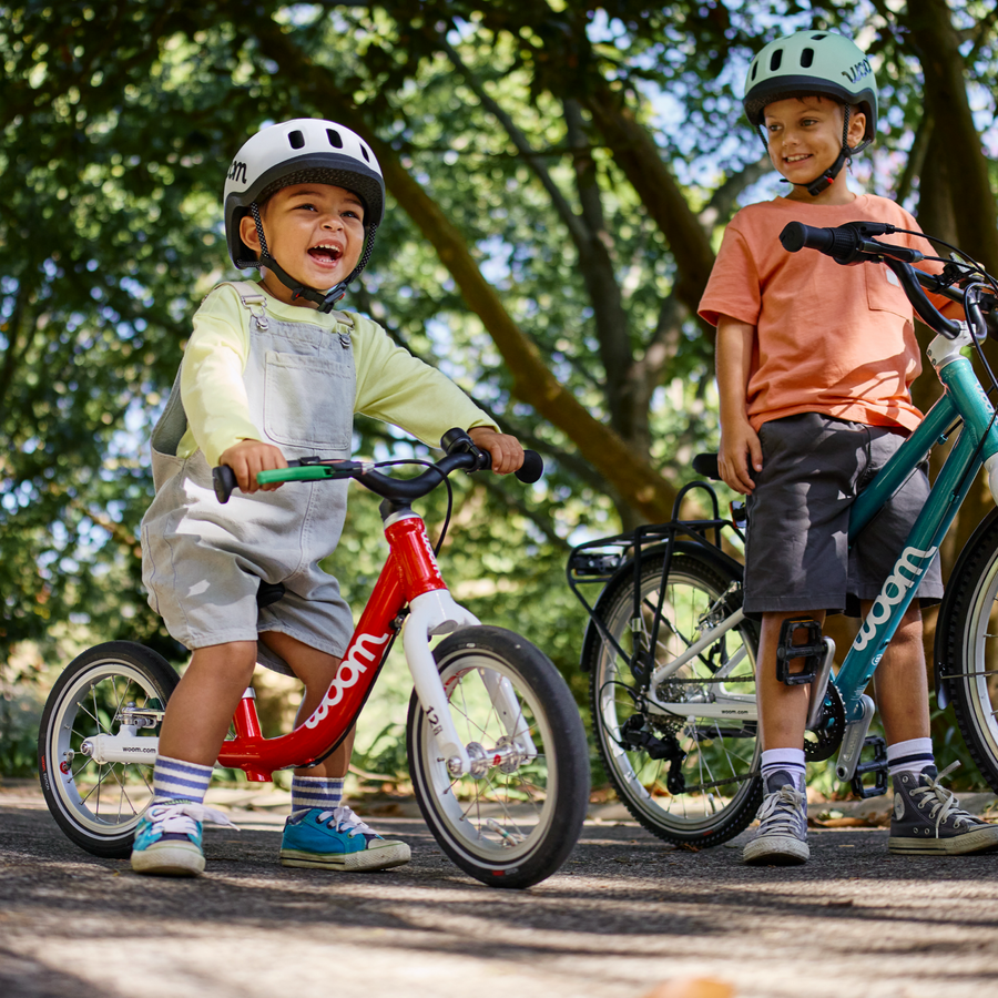 Two children on Woom balance bikes in a park setting with trees in the background.