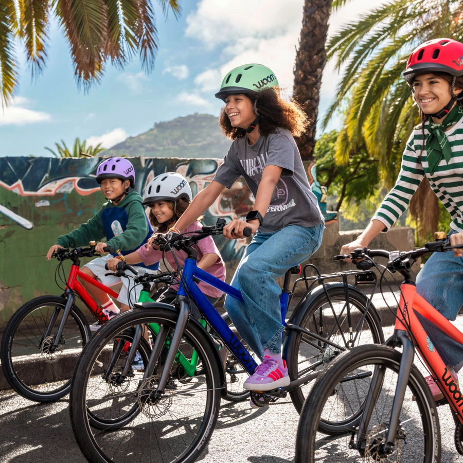Children riding Woom bicycles on a street with palm trees and a mural in the background