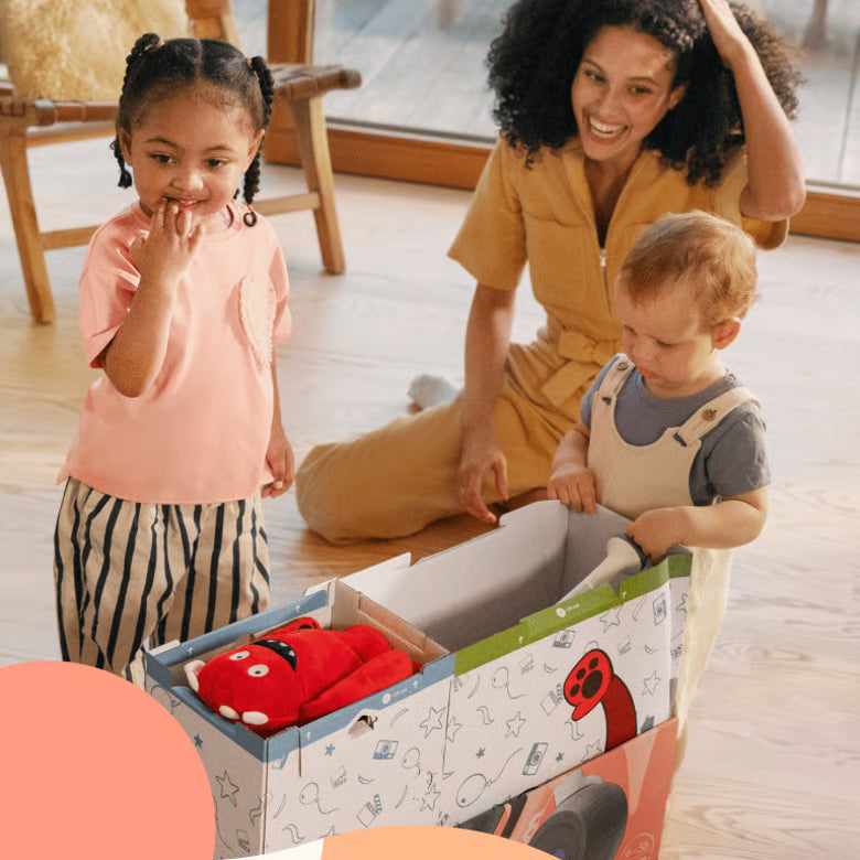 A woman and two children are unpacking a balance bike on a wooden floor.