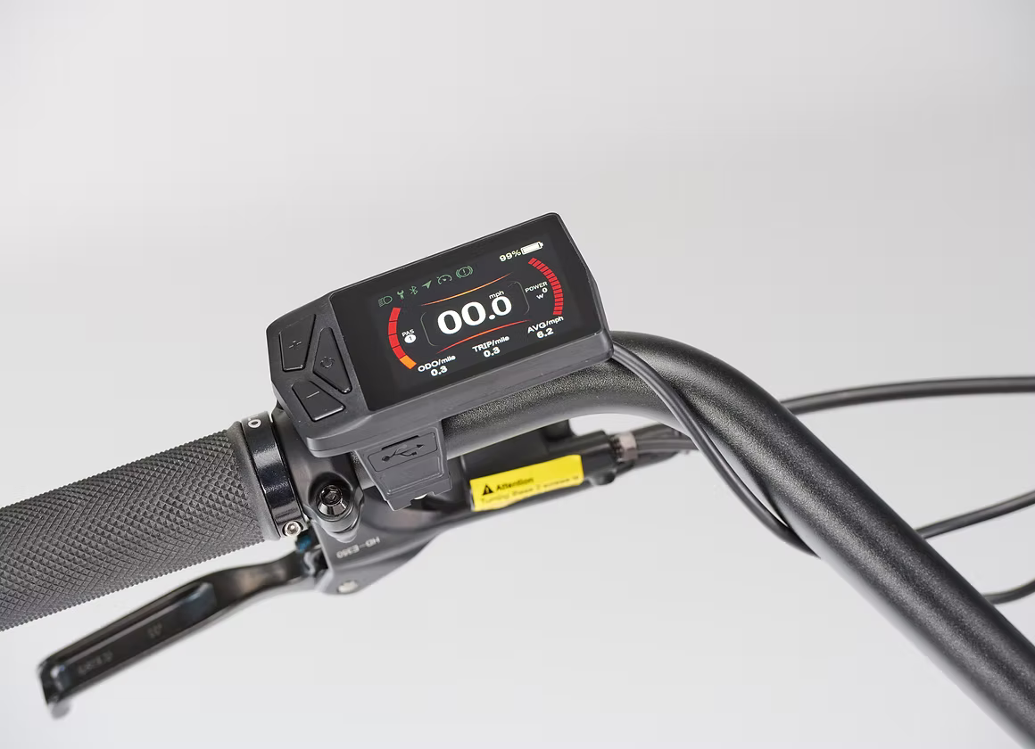 Close-up of a bicycle or e-bike handlebar with a digital display mounted on a black handlebar clamp, showing a speedometer-style screen and control buttons, photographed against a clean light gray background.