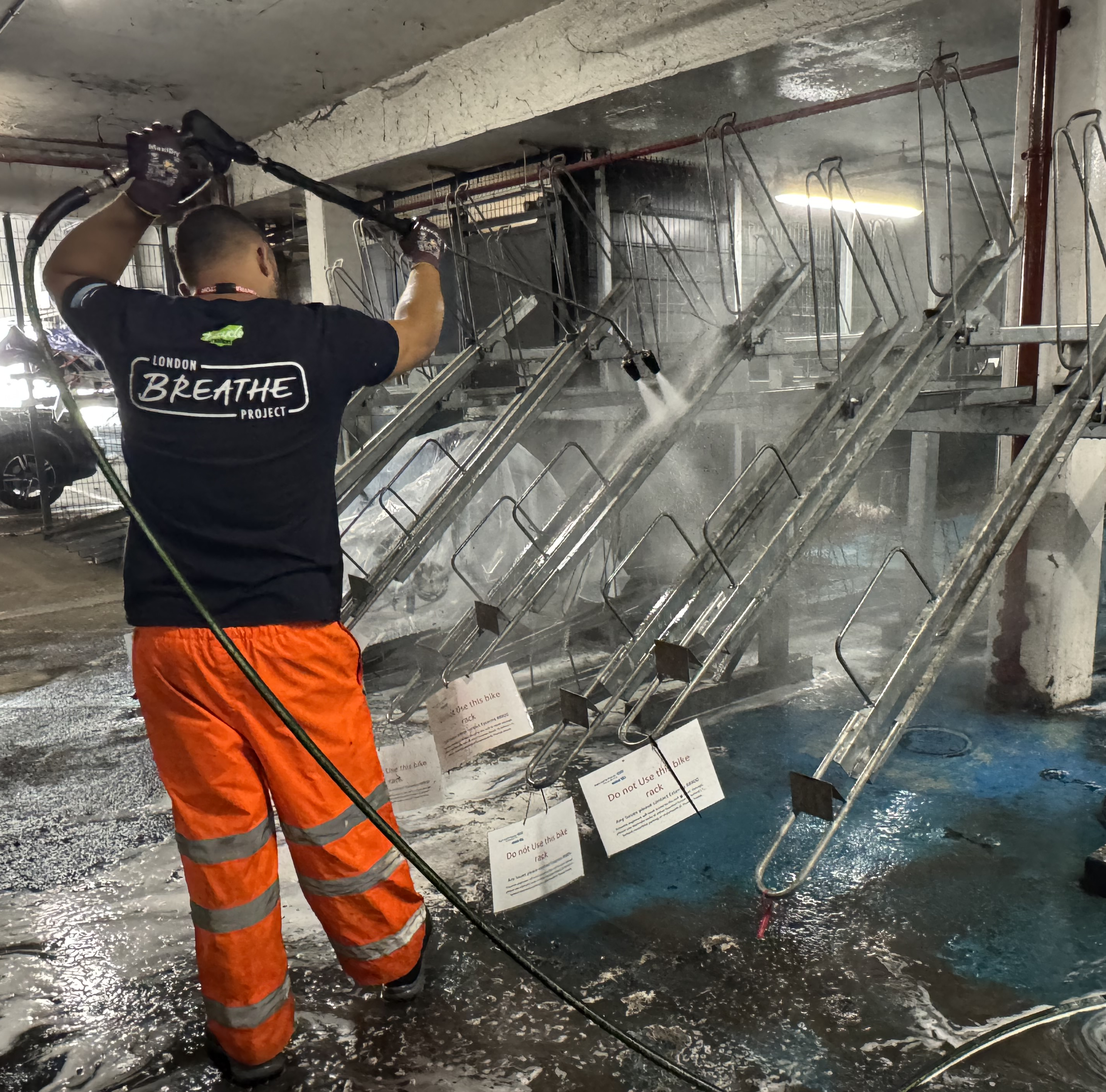 Person cleaning bicycle racks with a pressure washer wearing a 'Breathe London' t-shirt.