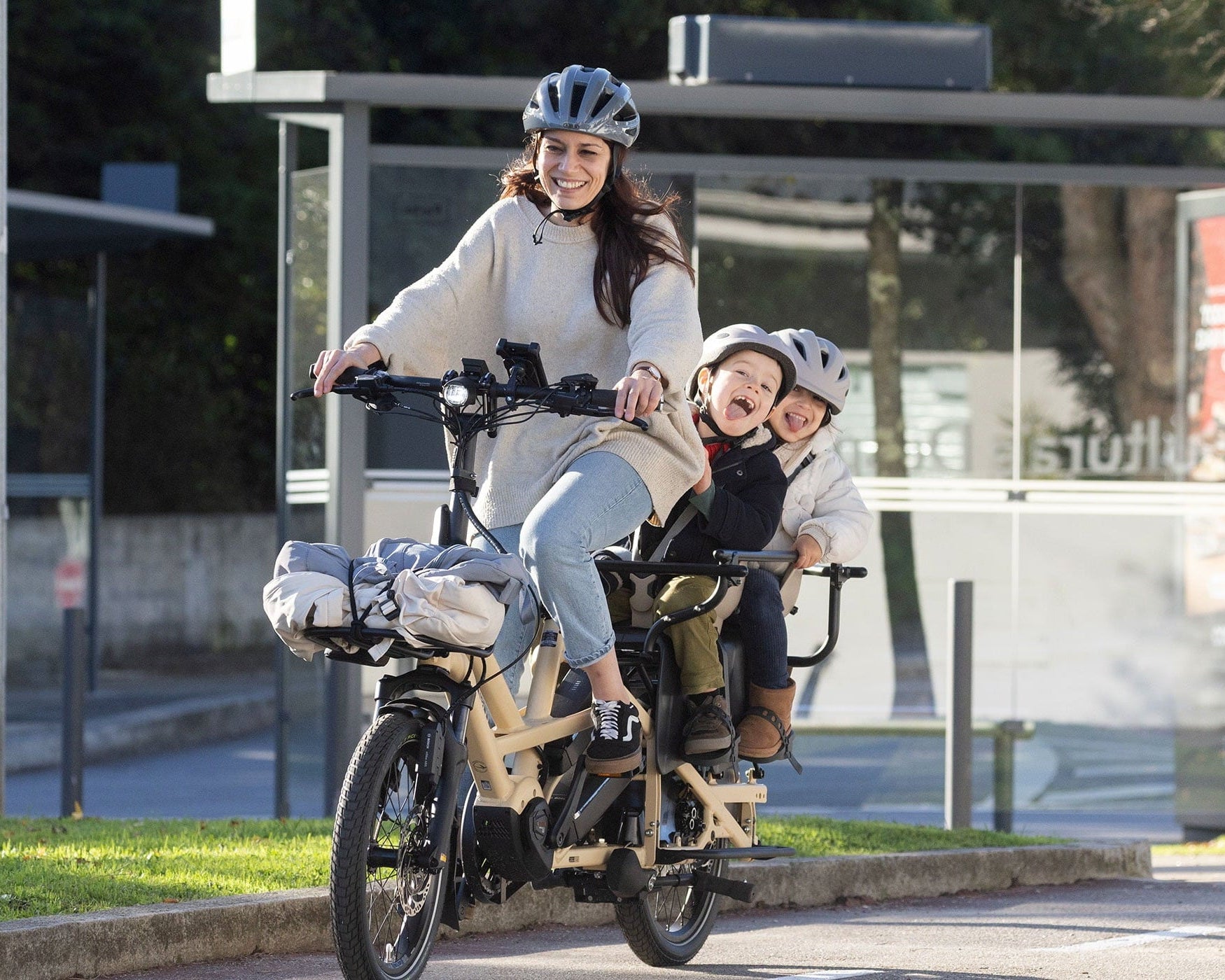 Woman riding a bicycle with two children in a bike trailer on a city street.